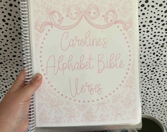 以下が含まれることがあります： ピンクの花柄と「Caroline's Alphabet Bible Verses」の文字が入った、スパイラル綴じのノート。表紙にはピンクのリボンと花のボーダーがあしらわれています。ノートは人の手で持たれています。