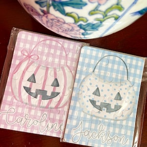 May include: Two small Halloween-themed jigsaw puzzles. One is pink gingham with a pumpkin design and the name "Caroline". The other is blue gingham with a pumpkin design and the name "Jackson". Both are in clear plastic.