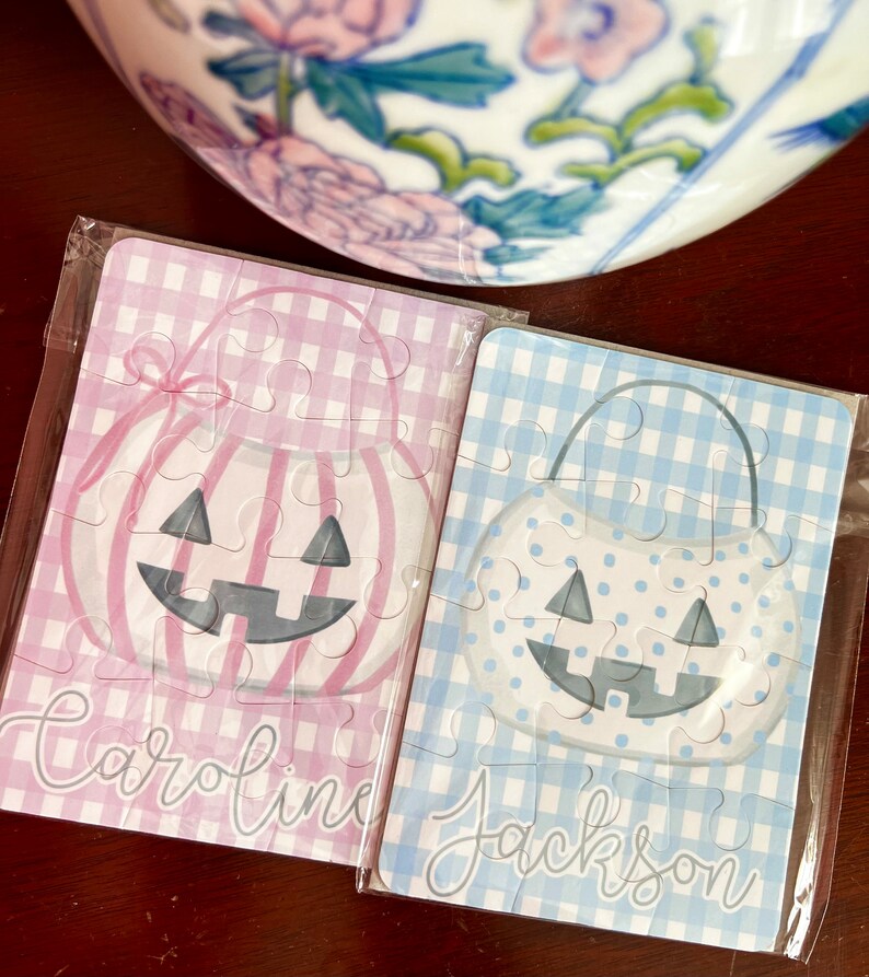 May include: Two small Halloween-themed jigsaw puzzles. One is pink gingham with a pumpkin design and the name "Caroline". The other is blue gingham with a pumpkin design and the name "Jackson". Both are in clear plastic.