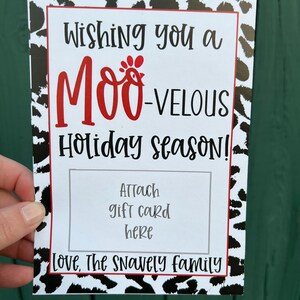 May include: A white card with black and white cow print design. The text "Wishing you a MOO-VELOUS Holiday season!" is in black with a red chicken graphic. The text "Attach gift card here" is in black. The text "Love, The Snavely family" is in black.