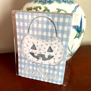 May include: A blue and white gingham patterned puzzle in the shape of a pumpkin, with a smiling face and the name "Jackson" in script. The puzzle is in a clear plastic wrapper.