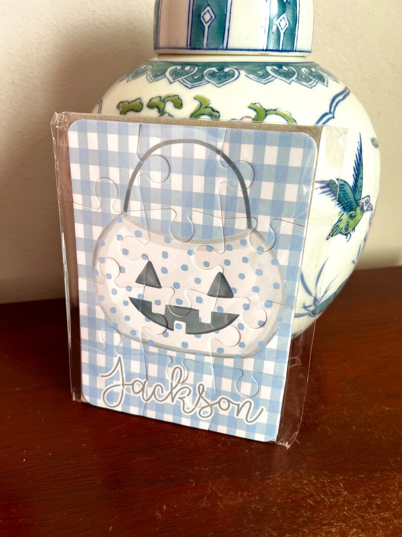 May include: A blue and white gingham patterned puzzle in the shape of a pumpkin, with a smiling face and the name "Jackson" in script. The puzzle is in a clear plastic wrapper.