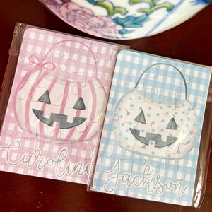 May include: Two small Halloween-themed jigsaw puzzles. One is pink gingham with a pumpkin design and the name "Caroline". The other is blue gingham with a pumpkin design and the name "Jackson". Both are in clear packaging.