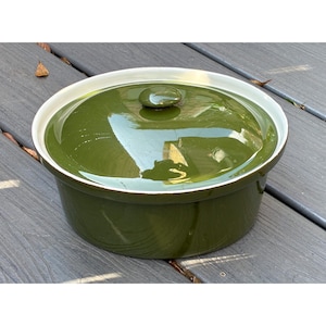 Puede incluir: Una fuente de horno redonda de cerámica en color verde oliva con tapa a juego. El recipiente presenta un acabado brillante y liso, un pomo sencillo en el centro de la tapa y un borde interior blanco que contrasta en la parte superior.