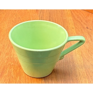 Puede incluir: Una taza de cerámica de color verde claro con un acabado liso y una abertura ancha. El exterior presenta tres anillos horizontales en relieve que rodean la parte central del cuerpo. Incluye un asa angular unida al lateral de la taza.