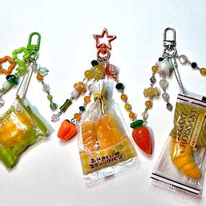 May include: Four food-themed keychains with decorative beads and charms. Each keychain features a miniature food item, including a melon bun, bread, a croissant, and a sandwich. The keychains have various colored clasps and charms.