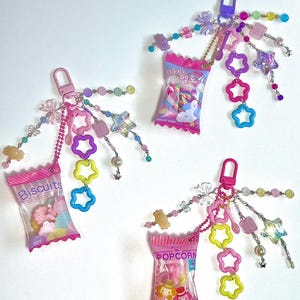 May include: Three pink and white candy bags with colorful charms attached. The bags are labeled "Biscuits", "Popcorn", and "Candy". The charms include stars, flowers, and beads.
