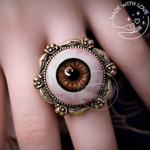 May include: A ring featuring a central realistic brown human eye cabochon with visible red veins, set within an antique brass-toned metal frame decorated with leaf-like motifs. The design includes a circular border around the eye and a textured finish.