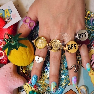 May include: A collection of rings including a pink plastic ring, a silver heart ring, a gold-toned locket ring with the text MY BUTT, a gold-toned ship wheel ring, two gold-toned BFF rings, and a wavy silver ring, all worn on fingers.