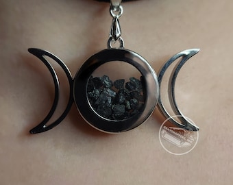 Puede incluir: Un collar con un colgante de plata en forma de triple luna con un círculo de vidrio transparente que contiene piedras negras. El colgante está en un choker negro.