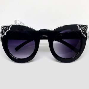 May include: Black cat-eye sunglasses with a spiderweb design on the temples. The lenses are a dark gray color.