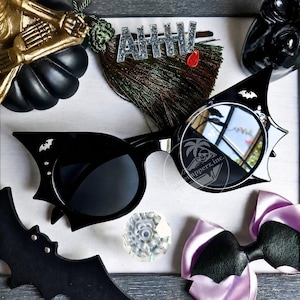 Puede incluir: Gafas de sol negras con un diseño de montura en forma de alas de murciélago que presenta pequeñas siluetas de murciélagos plateados en las esquinas exteriores. Las lentes son oscuras y circulares, con un sutil logotipo de marca de agua.