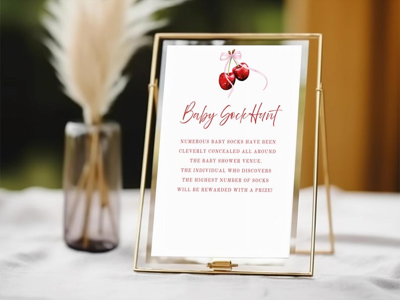 Op de afbeelding: Een wit bord met rode tekst in een gouden lijst die een "Baby Sokken Jacht" aankondigt. Het bord zegt: "Talrijke babysokken zijn slim verborgen in de hele baby shower locatie. De persoon die de meeste sokken vindt, wordt beloond met een prijs!" Een roze lint met twee rode kersen bevindt zich bovenaan het bord.