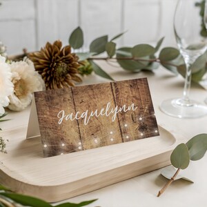 May include: A folded rectangular place card featuring a rustic brown wood grain pattern with a string of white fairy lights printed across the bottom. The name Jacquelynn is written in a white cursive script font across the center of the card.