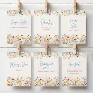 Puede incluir: Seis tarjetas rectangulares blancas colgadas de una cuerda con pinzas de madera. Cada tarjeta presenta un borde de flores silvestres coloridas en la parte superior e inferior, con texto azul y letras rojas para señalización de eventos.