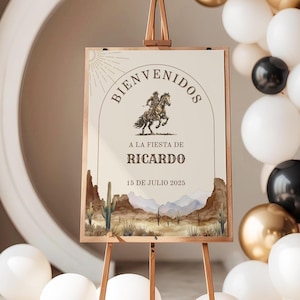 May include: A rectangular welcome sign on a wooden easel featuring a desert landscape with cacti and mountains at the bottom. The text reads BIENVENIDOS A LA FIESTA DE RICARDO 15 DE JULIO 2025, with a central illustration of a person riding a horse.