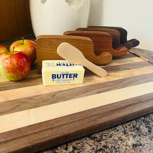 May include: A rectangular striped wooden cutting board with three upright wooden butter dishes, a light-colored wooden butter spreader, a stick of salted butter labeled "BUTTER 1 FIRST QUALITY 1 NET WT 4 OZ (113g)", and two red and yellow apples.