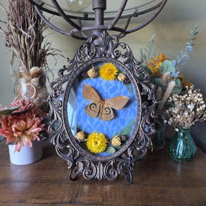 May include: An antique-style, oval frame with a preserved moth specimen. The moth is brown and tan, set against a blue patterned background. Yellow flowers and greenery surround the moth. The frame is supported by a black stand.