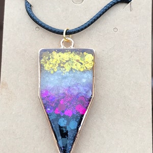 May include: A gold pendant necklace with a black cord. The pendant is a triangle shape with a clear resin filled with glitter in yellow, white, purple, and black colors. The pendant has a gold trim. The tag below the necklace reads "Handmade with love."