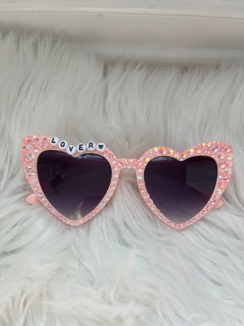 May include: Pink heart-shaped sunglasses with rhinestones and the word "LOVER" spelled out in black letters on the frame.