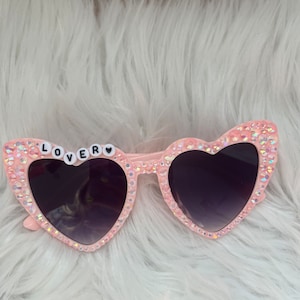 May include: Pink heart-shaped sunglasses with rhinestones and the word "LOVER" spelled out in white letters on the left lens.
