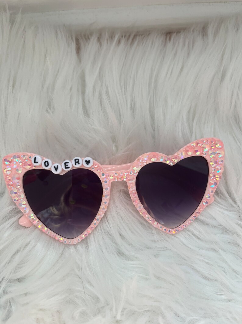 May include: Pink heart-shaped sunglasses with rhinestones and the word "LOVER" spelled out in white letters on the left lens.