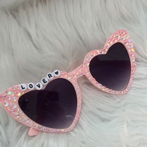 May include: Pink heart-shaped sunglasses with rhinestones and the word "LOVER" spelled out in white letters on the left lens.