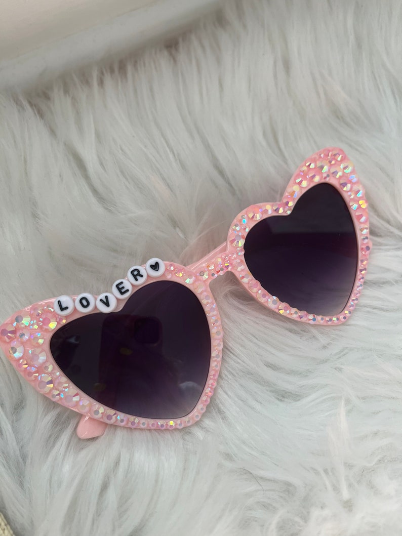May include: Pink heart-shaped sunglasses with rhinestones and the word "LOVER" spelled out in white letters on the left lens.