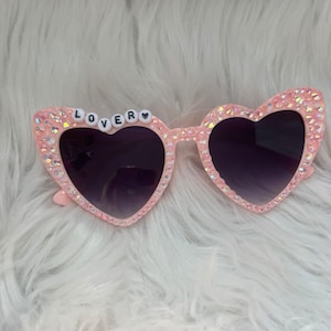 May include: Pink heart-shaped sunglasses with rhinestones and the word "LOVER" spelled out in white letters on the frame.