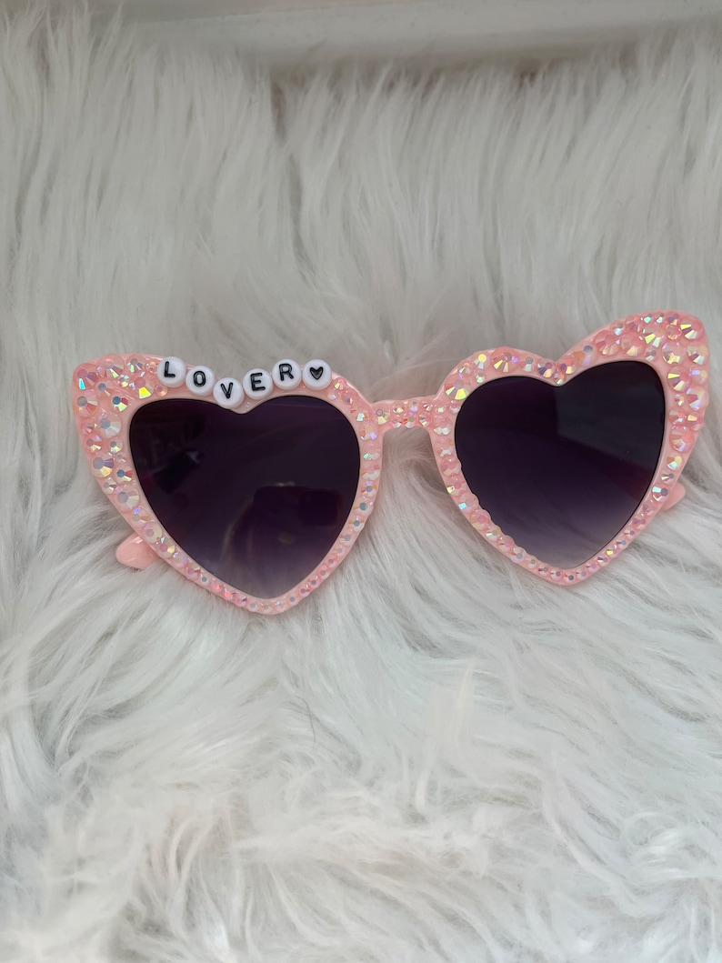 May include: Pink heart-shaped sunglasses with rhinestones and the word "LOVER" spelled out in white letters on the frame.