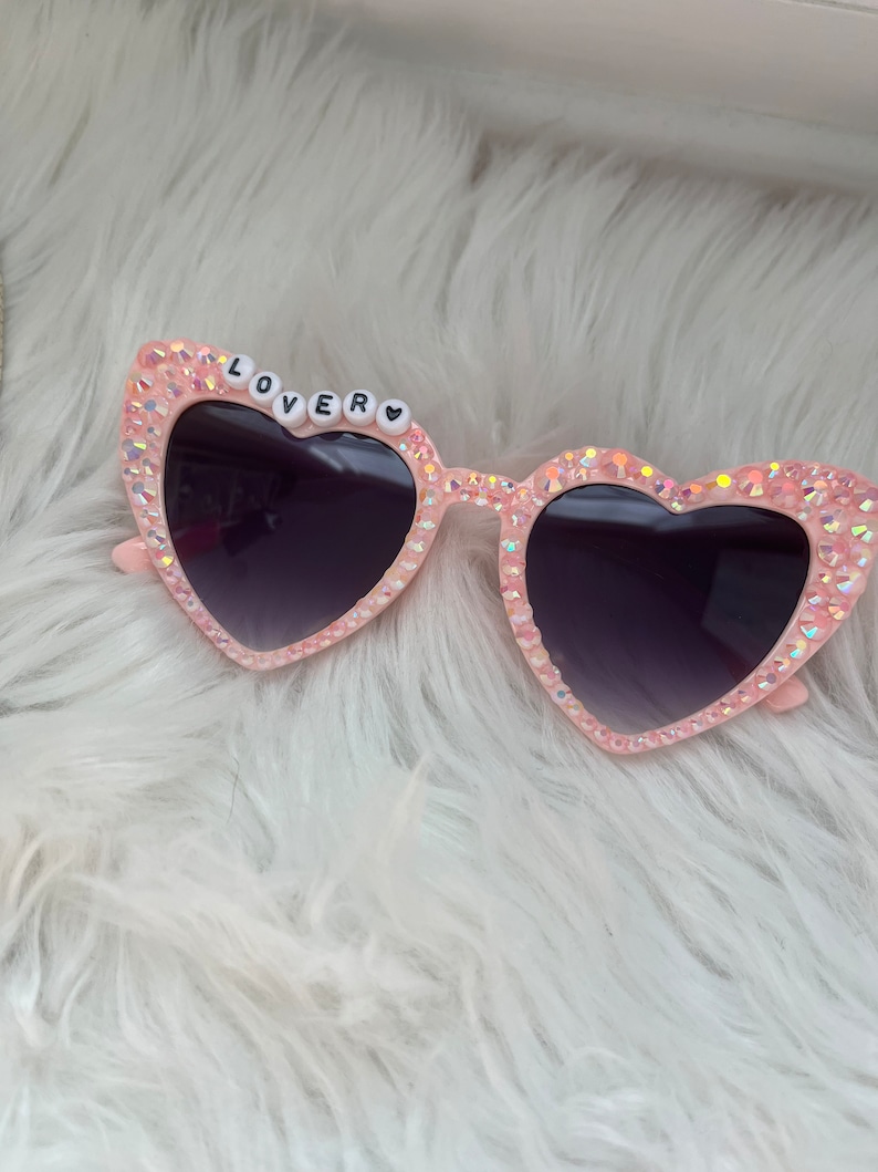 May include: Pink heart-shaped sunglasses with rhinestones and the word "LOVER" spelled out in white beads.
