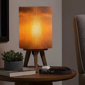 May include: A wooden table lamp with a cylindrical, grooved shade. The lamp is supported by a tripod base. The warm light from the lamp brightens the room. A small potted plant and a tray with accessories are on the table.