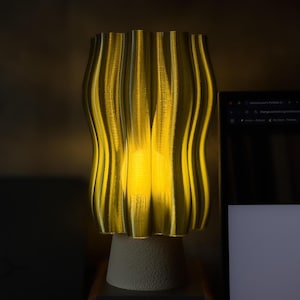 Könnte beinhalten: Eine gelbe Lampe mit wellenförmigem Muster und einem strukturierten, cremefarbenen Sockel. Die Lampe strahlt ein warmes, goldenes Leuchten aus und erzeugt ein weiches Umgebungslicht. Das Design weist vertikale, geschwungene Linien auf und wirkt modern und künstlerisch.
