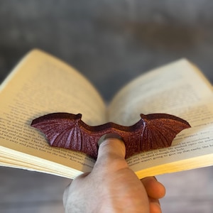 May include: A burgundy bat-wing bookmark is placed in an open book. The bookmark, with a textured finish, is held by a finger. The book's pages are yellowed, and text is visible. The bookmark is a unique accessory.