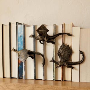 May include: Three bronze-coloured dragon figurines attached to the spines of a row of books. Each dragon is depicted in a flying pose with detailed wings, textured scales, and long tails, positioned horizontally across the book spines.