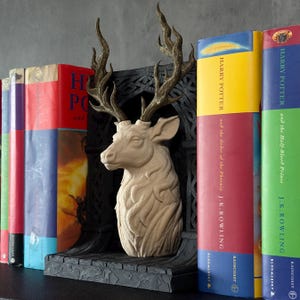 May include: A beige deer head bookend with bronze-colored antlers stands between colorful Harry Potter books. The bookend has a detailed, textured design and sits on a dark gray base. The books have titles by J.K. Rowling.