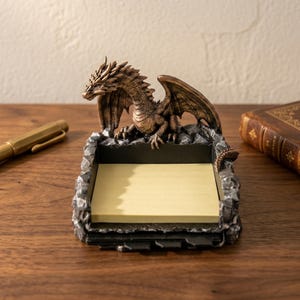May include: A bronze dragon ornament sits on a memo pad holder. The dragon has detailed wings and scales, with a gray and black base. The memo pad is filled with yellow sticky notes. A gold pen and a leather-bound book are in the background.