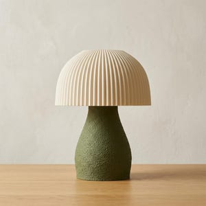 May include: A mushroom-shaped lamp with a textured, olive green base and a cream-coloured, ribbed lampshade. The lamp is placed on a wooden surface, providing a unique lighting solution for any room. The lamp's height is approximately 25 cm.