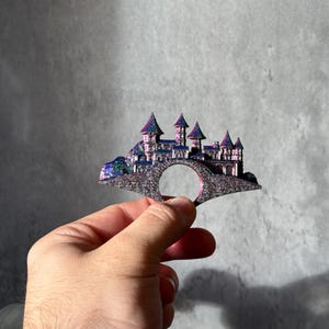 May include: A fantasy-themed ring featuring a castle design with multiple towers and a bridge. The ring has a glittery, iridescent finish with shades of purple, blue, and silver. The ring is held in a hand against a gray background.