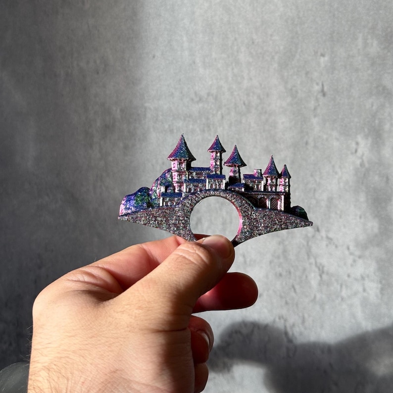 May include: A fantasy-themed ring featuring a castle design with multiple towers and a bridge. The ring has a glittery, iridescent finish with shades of purple, blue, and silver. The ring is held in a hand against a gray background.