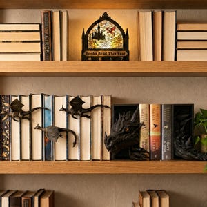 May include: A decorative arched light box with a stained glass dragon and castle design, featuring the text "Books Read This Year" and numerical counters. Below, three small bronze-coloured dragon figures are mounted on book spines.