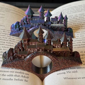 May include: A detailed book page holder shaped like a castle, with a bridge and multiple towers. The castle is a mix of brown and iridescent colors, and is designed to hold a book open. The holder is made of a sturdy material.