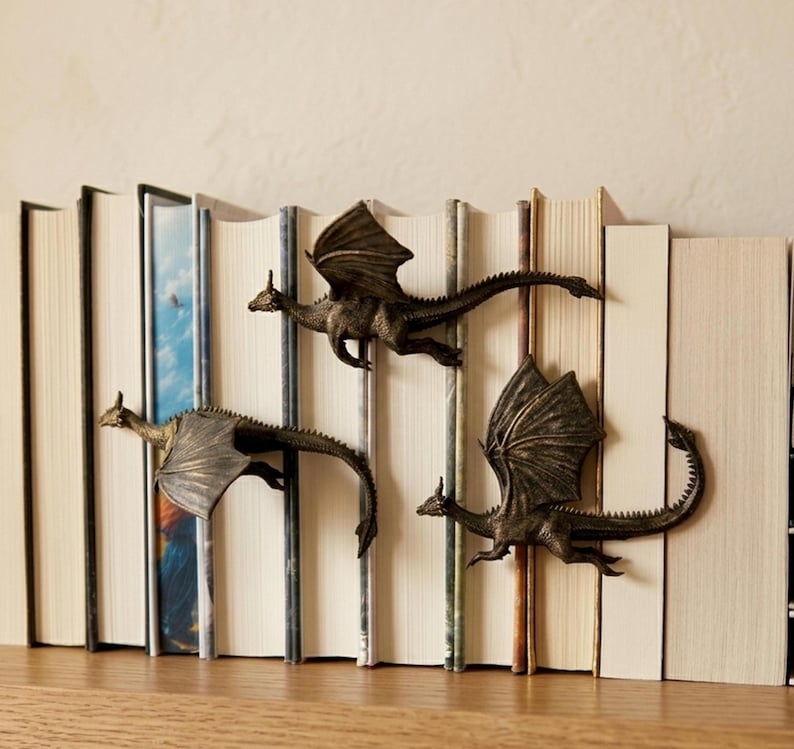 May include: Three bronze-coloured dragon figurines attached to the spines of a row of books. Each dragon is depicted in a flying pose with detailed wings, textured scales, and long tails, positioned horizontally across the book spines.