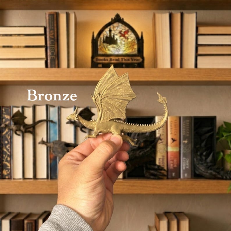 May include: A bronze-coloured dragon figurine with detailed wings, a long spiked tail, and a textured body. The dragon is held in a hand against a background of bookshelves. The text "Bronze" is visible on the left side of the image.