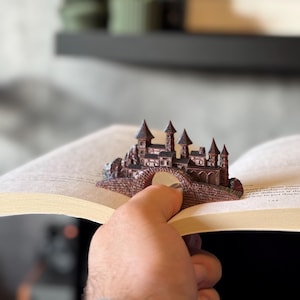 May include: A detailed, miniature castle sculpture in shades of brown and bronze, resting on a bridge. The castle features multiple turrets and architectural details. The sculpture is placed on the open pages of a book.