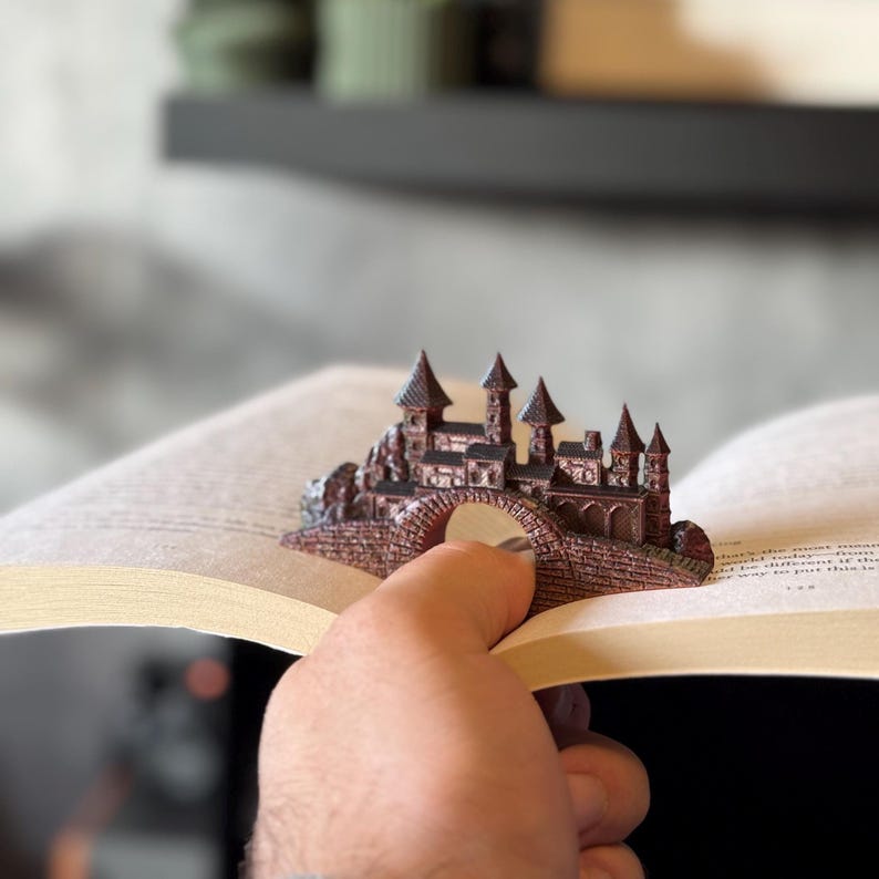 May include: A detailed, miniature castle sculpture in shades of brown and bronze, resting on a bridge. The castle features multiple turrets and architectural details. The sculpture is placed on the open pages of a book.