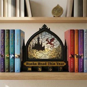 Peut inclure: Un serre-livres noir décoratif avec un motif de vitrail représentant un château et un dragon, avec le texte "Books Read This Year". Flanqué de livres Harry Potter de différentes couleurs.