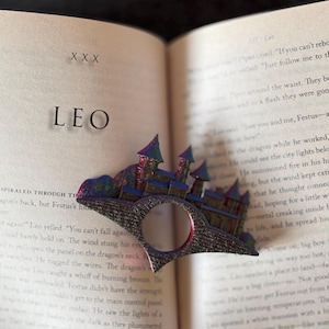 May include: A detailed bookmark shaped like a castle, with a bridge and towers. The bookmark is made of a dark metallic material with blue, purple, and pink accents. The word "LEO" is visible on the open book.