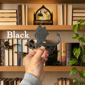 May include: A matte black dragon figurine with detailed wings, a long spiked tail, and a slender body. The dragon is held in a position that displays its full profile, including the textured wing membranes and the sharp, pointed tail tip.