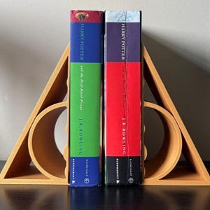 May include: Two "Harry Potter" books, "and the Half-Blood Prince" and "and the Deathly Hallows", are held by a wooden bookend. The bookend is light brown and shaped like a triangle with a circle cut out. The books have colourful spines.